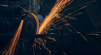 Close up of worker in special black suit working with angle grinder and grinding metal with large yellow sparks. Concept of polishing metal with special equipment in dark room.